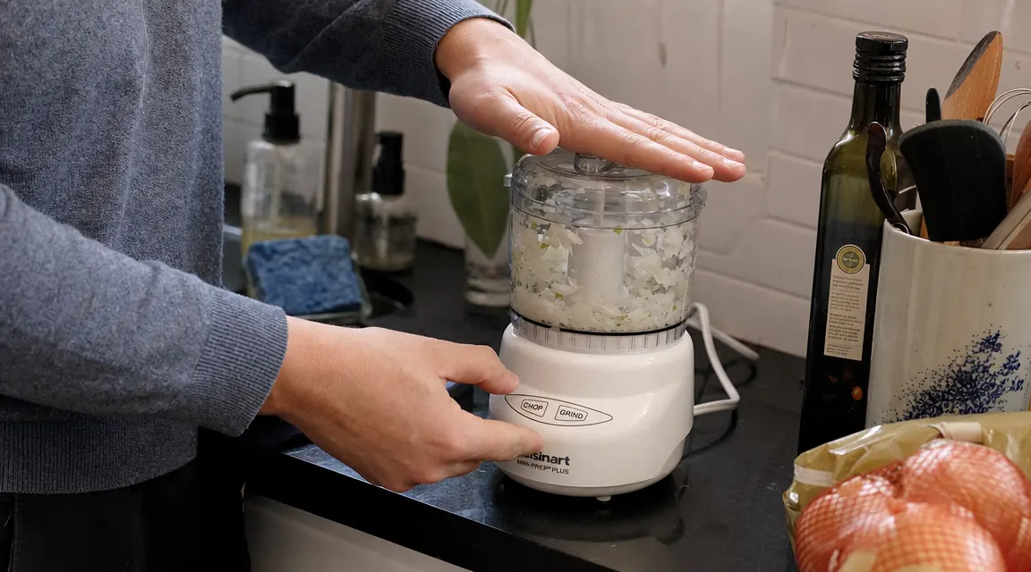 Hand pressing down the lid of the Cuisinart Mini-Prep Plus to pulse chopped onions, kitchen counter in background